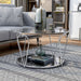 Right angled modern round chrome steel coffee table with open teardrop shape legs, a decorated tempered glass top and mirror open bottom shelf on a rug in front of a sofa.