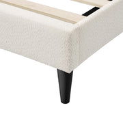 Left facing close up of the footboard corner and support leg of a modern white boucle upholstered queen bed on a white background