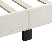 Left facing close up of the side rail, slats, and support leg of a modern white boucle upholstered queen bed on a white background