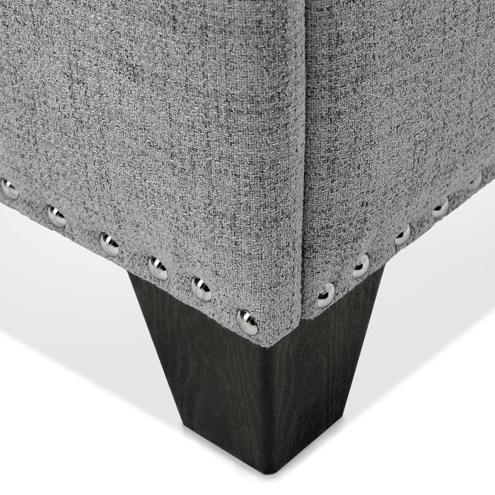 Right facing close up of the nailhead trim and leg of a modern grey upholstered storage bench with a lift top on a white background