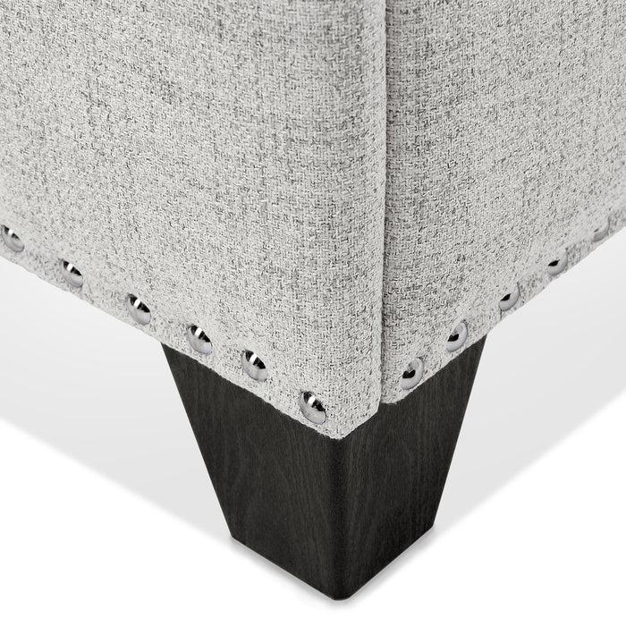 Right facing close up of the nailhead trim and leg of a modern off white upholstered storage bench with a lift top on a white background