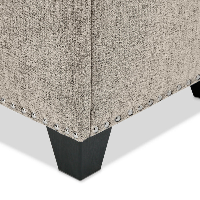 Right facing close up of the nailhead trim and leg of a modern light brown upholstered storage bench with a lift top on a white background