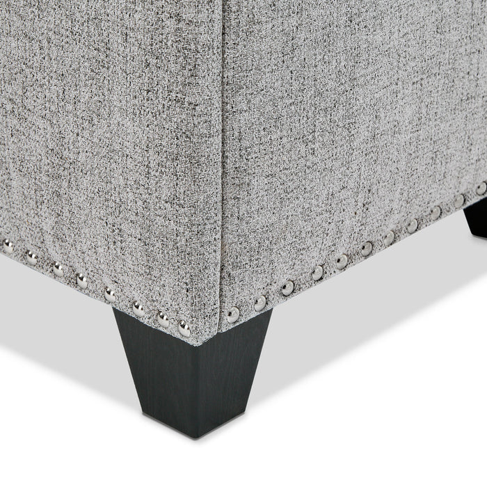 Right facing close up of the nailhead trim and leg of a modern light grey upholstered storage bench with a lift top on a white background