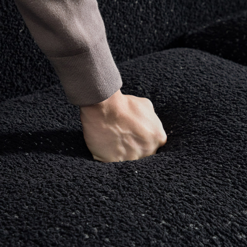 Right facing close up of a person's fist pressing into the seat cushion of a contemporary black metallic boucle shelter loveseat