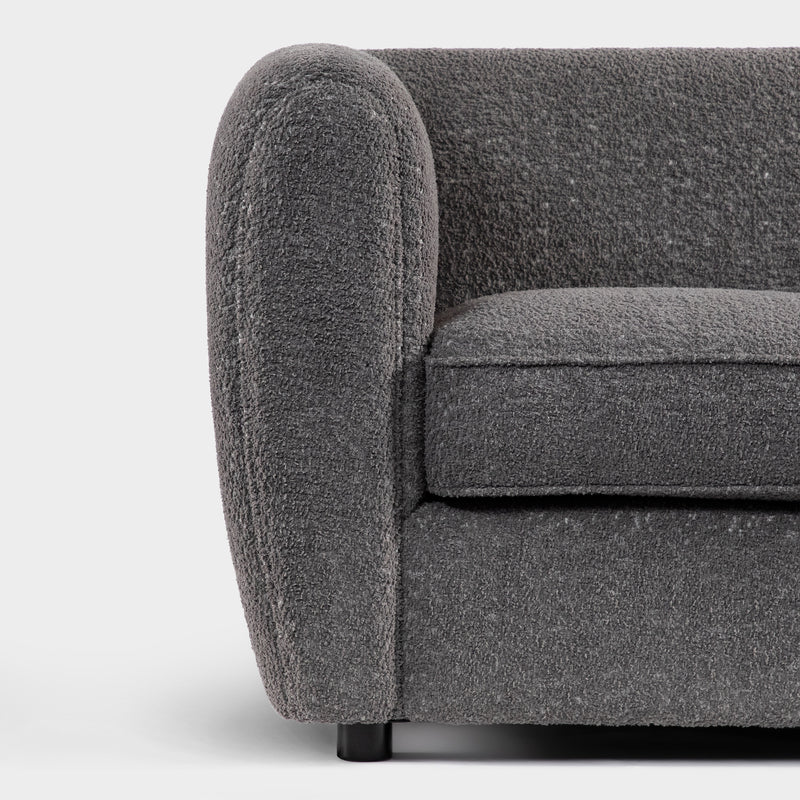 Front facing close up of the rounded armrest and leg of a contemporary charcoal gray metallic boucle shelter sofa on a white background