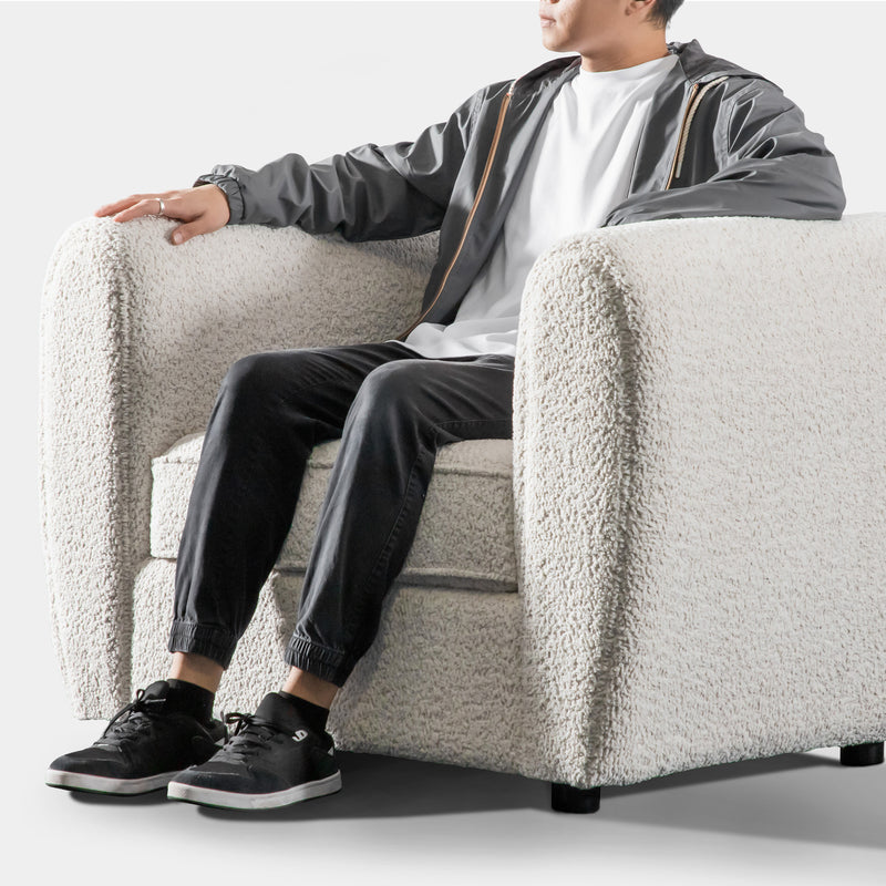 Left facing close up of a person seated on a contemporary off-white metallic boucle shelter armchair on a white background