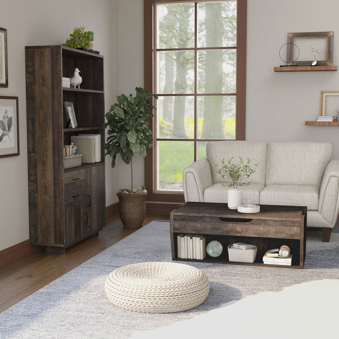 Side view of a reclaimed oak storage coffee table and bookcase set in a modern farmhouse style living room. The chest-like coffee table, in front of the beige loveseat, features an open shelf on the base frame displaying books and a basket. Three spacious shelves on the bookcase display more books and decor. An indoor tree cushions the corner between the bookcase and loveseat, while a round rattan floor ottoman sits in the foreground of the coffee table.