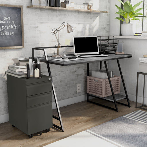 Right-angled urban grey computer desk and file pedestal set in a home office. A laptop, lamp, and paper tray fill the desktop, while the lower open shelf of the desk holds a basket of textbooks. The gunmetal 3-drawer file pedestal keeps an accessory holder.
