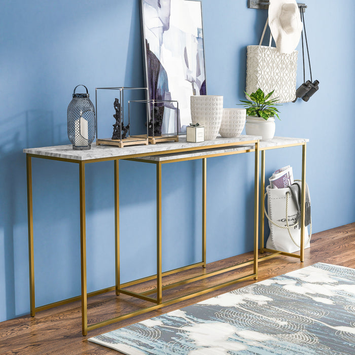 Right-angled faux white marble nesting tables with gold-tone frames against a light cobalt blue wall. A modern blue painting, vases, and decor adorn the tabletops, while bags are stored next to the entryway tables.