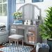 Left-angled silver vanity set in a blue themed youth bedroom. Make-up adorns the vanity table as the silver set mingles with the blue accents of the room. A blue curtain hangs to the left, two blue art frames hang just above the mirror, and the stool sits on a blue patterned rug. To the right blue linen drapes over a grey wicker basket and a plant sits in a blue pot.