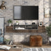 Front-facing reclaimed oak wall-mounted TV console in an urban living room. A chicken wire basket of books and a cognac leather ottoman sit under the floating TV console. A VCR, stereo, x-box, and controller sit on its open shelves. A TV is mounted just above the console.
