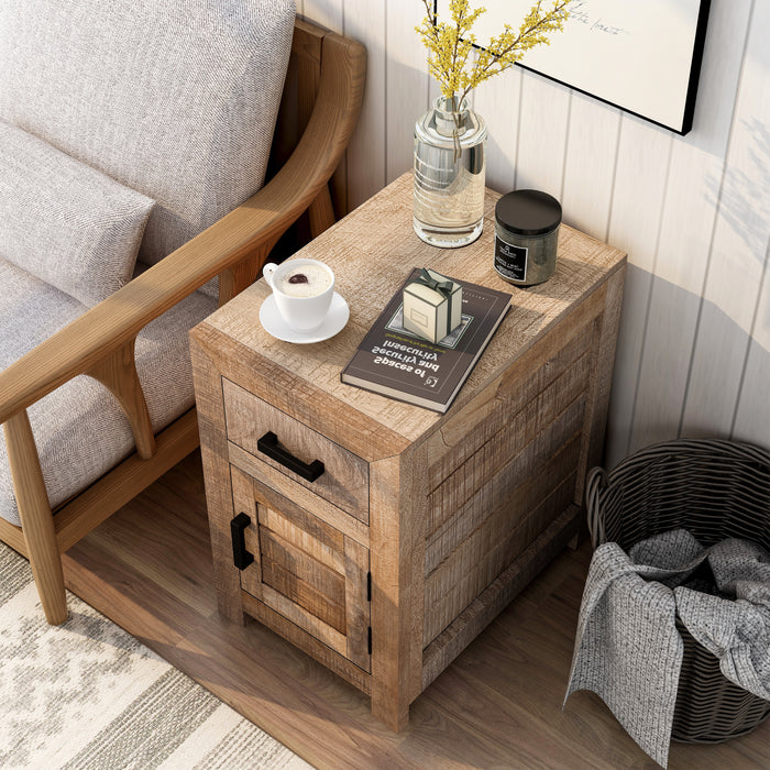 Top view of a solid mango wood side table in a beach house. The unfinished sawblade design and black bar pulls add a casual look to the shiplap white wall. A black frame hangs above the end table while a black basket sits to its right. A vase of yellow flowers, a book, and a macchiato sit on the table next to a sofa.