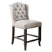 Right-angled beige counter-height chair against a white background. The button-tufted wingback chair and seat are accented in nailhead trim. An antique black leg frame offers footrests for comfort and rustic style.