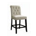 Right-angled beige counter height chair against a white background. The scrolled back is adorned with button-tufting and a welted trim. Its antique black frame features footrests.