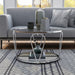 Front-facing modern round chrome steel coffee table with open teardrop shape legs, a decorated tempered glass top and mirror open bottom shelf on a rug in front of a sofa.