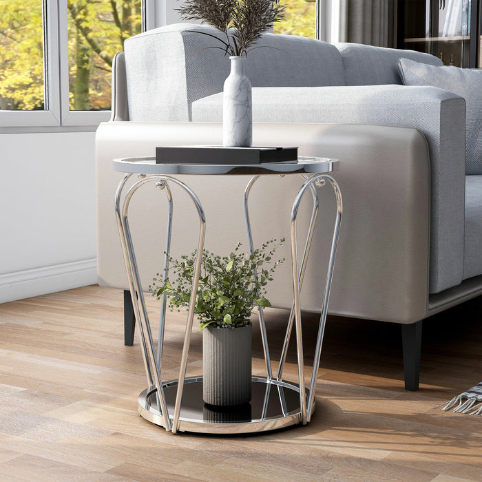 Left angled modern round chrome end table with open teardrop shape steel legs, a clear tempered glass top, and mirror open bottom shelf decorated with plant and books next to a sofa.