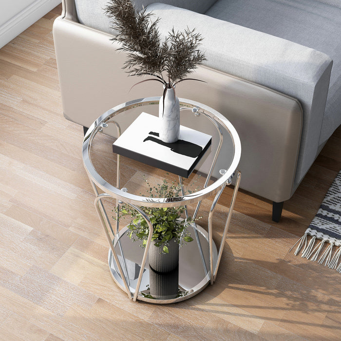 Left angled top-down modern round chrome end table with open teardrop shape steel legs, a clear tempered glass top, and mirror open bottom shelf decorated with plant and books next to a sofa.