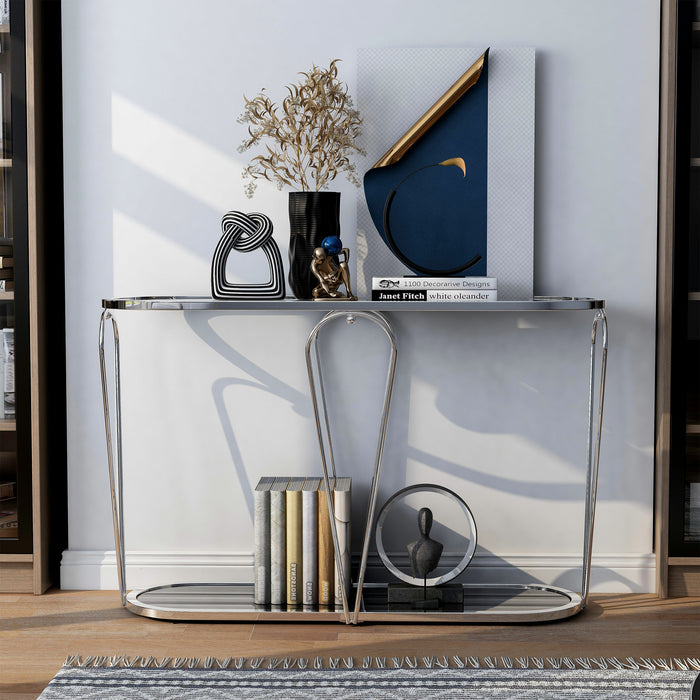 Front-facing modern chrome console table with open teardrop shape steel legs, rounded tempered glass top, and mirror open bottom shelf with books and decor against a wall.