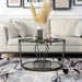 Front-facing modern round black nickel coffee table with open teardrop shape steel legs, a gray tempered glass top, and mirror open bottom shelf decorated with accessories on a rug in front of a sofa.
