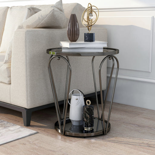 Left angled modern round black nickel end table with open teardrop shape steel legs, a gray tempered glass top, and mirror open bottom shelf decorated with books and accessories next to a sofa.