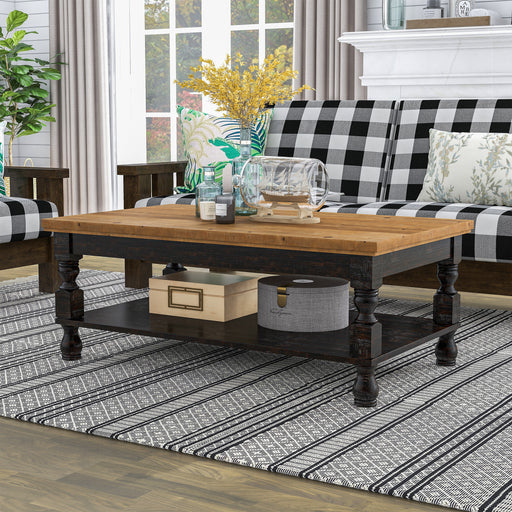 Left angled rustic antique black wood coffee table with oak tabletop and open lower shelf decorated with accessories in a living room.