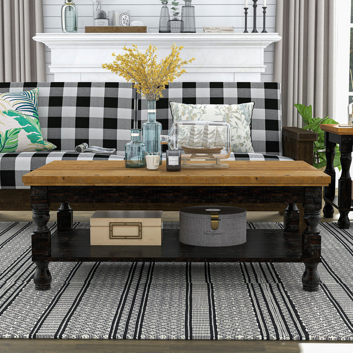 Front-facing rustic antique black wood coffee table with oak tabletop and open lower shelf decorated with accessories in front of a sofa.