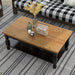Left angled top-down rustic antique black wood coffee table with oak tabletop decorated with accessories on a rug in front of a sofa.