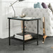 Left angled modern industrial black steel end table with tempered white marble glass top, slender steel legs and perforated open metal shelf decorated next to a sofa.