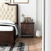 Front-facing cherry nightstand in a traditional bedroom. From the doorway view, it sits to the right of a cherry framed and beige headboard upholstered bed. 