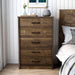 Front-facing rustic walnut 5-drawer chest in a farmhouse-style bedroom. The case good holds a tray with a candle, incense, and a small plant. The rustic bed on the right is adorned with neutral bedding while a gold frame hangs above the chest on the clean white shiplap wall.