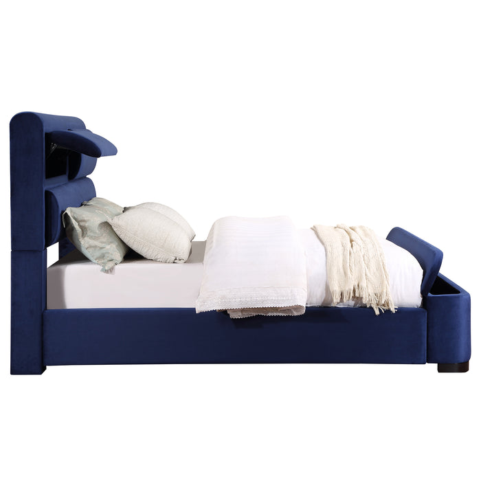 Front-facing side view of a modern glam navy blue upholstered bed with head and footboard storage and linens on a white background