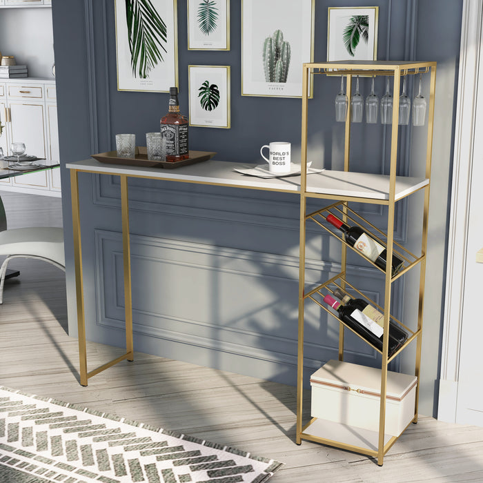 Left-angled high gloss white and gold bar table in a glam kitchen. A built-in tier accompanies the white tabletop. Two shelves sandwich three hanging stemware racks with martini glasses and 8-bottle wine racks. Two glasses or red wine sit on the tabletop bar along with two books. Luxury gold bar chairs match the energy of the furnishing.