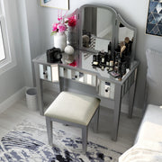 Top view of a silver vanity set in a modern light grey bedroom. Creams, cosmetics, a camera, and a flower vase sit on the mirrored tabletop. Its positioned next to grey bedding on the right, while a light grey trash can accompanies its left. The light grey wood floors are adorned with an abstract blue rug and the light grey walls behind the vanity table are decked with modern blue wall art.