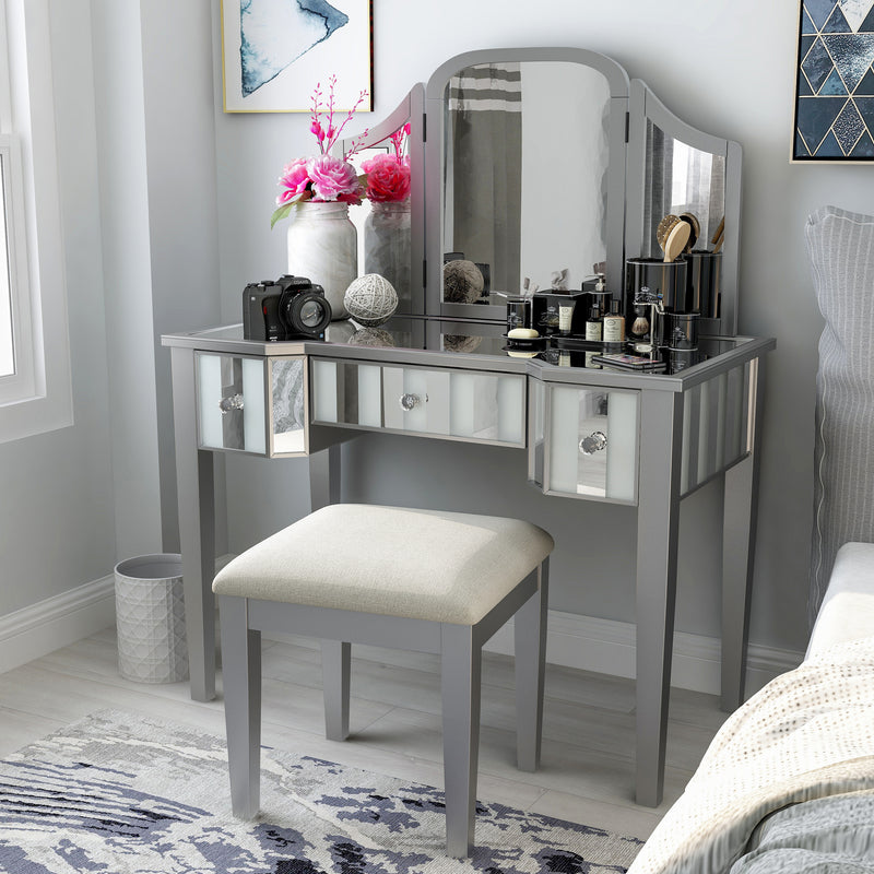Left-angled silver vanity set in a modern light grey bedroom. Creams, cosmetics, a camera, and a flower vase sit on the mirrored tabletop. Its positioned next to grey bedding on the right, while a light grey trash can accompanies its left. The light grey wood floors are adorned with an abstract blue rug and the light grey walls behind the vanity table are decked with modern blue wall art.