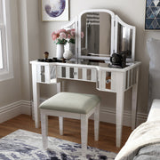 Left-angled white vanity set in a modern bedroom. Creams, cosmetics, a camera, and a flower vase sit on the mirrored tabletop. Its positioned next to grey bedding on the right, while a white trash can accompanies its left. An abstract blue rug adorns the floor and modern blue wall art deck the wall behind the vanity table.