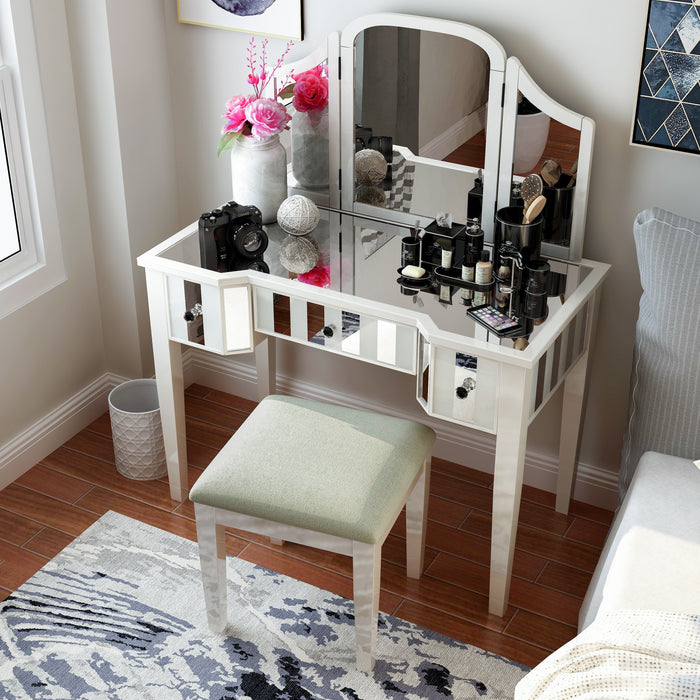 Top view of a white vanity set in a modern bedroom. Creams, cosmetics, a camera, and a flower vase sit on the mirrored tabletop. Its positioned next to grey bedding on the right, while a white trash can accompanies its left. An abstract blue rug adorns the floor and modern blue wall art deck the wall behind the vanity table.