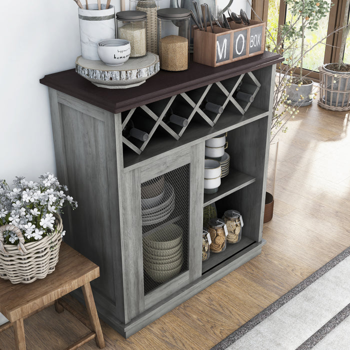 Top view of a vintage grey oak wine buffet against a white wall. Baking supplies present themselves on the tabletop while the lattice wine rack holds four bottles. On the open shelves are dishes and cookie jars. To the left of the server is a stool holding a basket of flowers.