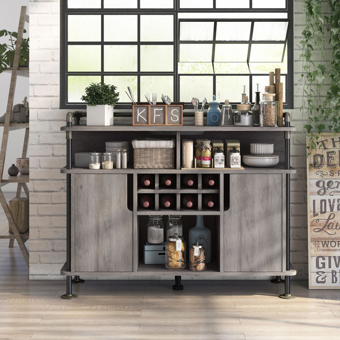Front-facing vintage grey oak wine cabinet in an urban loft. It sits on a taupe wood floor against a white brick wall, under a black-framed window. The pipe-accented lipped top displays silverware while the open shelves store other kitchen supplies. The wine rack holds 8-bottles. 