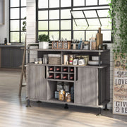 Left-angled vintage grey oak wine cabinet in an urban loft. It sits on a taupe wood floor against a white brick wall, under a black-framed window. The pipe-accented lipped top displays silverware while the open shelves store other kitchen supplies. The wine rack holds 8-bottles. 
